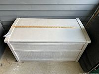 Top and side perspective of the light gray/taupe Suncast outdoor storage box with a 48-inch measuring stick on top showing approximate length.
