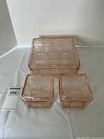 Photo showing all three pink Depression glass refrigerator dishes with matching lids on a white surface, exhibiting the geometric plaid pattern and square shapes.