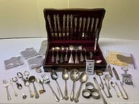 Full lot overview showing the wooden case with burgundy velvet lining holding an assortment of silver-plated flatware including forks, spoons, specialty serving utensils, napkin rings, and a corkscrew.