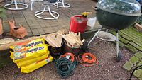Overall lot showing Weber charcoal grill, Duraflame logs, kindling wood in bins, gas can, electric cords coiled on ground, and grill brush.
