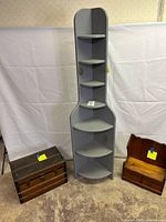 Tall grey corner shelf with six curved shelves between a wooden trunk and a small wooden wall shelf with drawers, all on concrete floor with white fabric backdrop