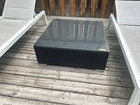 Square resin wicker coffee table with glass top between two lounge chairs on wooden deck