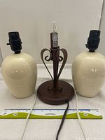 Photo showing the two cream-colored ceramic bedroom lamps without shades and the brown metal electric candle lamp with bulb all placed next to each other on a white surface.