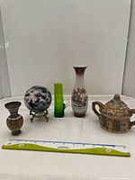 Front view of 5 assorted decorative items including copper pot, glass ball on holder, tall green glass, painted squirrel vase, and ceramic cottage container with lid, all placed on white cloth with ruler for scale.