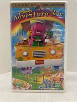 Front view of the VHS plastic case showing colorful artwork of Barney driving a yellow bus with Baby Bop and others, titled 'Barney's Adventure Bus' from the Classic Collection, 1997 release