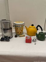 Coffee and tea items including the ceramic coffee container with scoop, two metal teapots (yellow and green), and vintage tea infuser on white fabric background