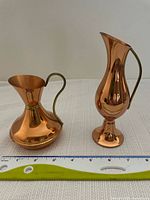 Both copper vases with brass handles side by side on a white surface with a measuring tape showing sizes