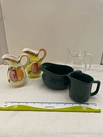 Five assorted sauce or juice containers: two ceramic fruit decorated pitchers, one clear glass pitcher, one dark green ceramic pitcher, and one dark green wide open jug/container.