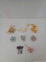 Photo showing 8 vintage brooches and a flower-combed hair accessory arranged on a neutral background. Includes various floral designs in gold-tone and silver-tone metals, accented with colored stones and pearls.