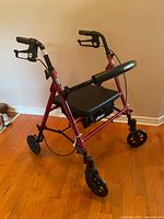 Red Drive walker with 4 wheels, hand brakes, padded seat and storage underneath on hardwood floor.
