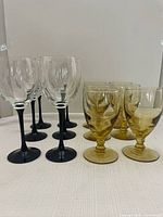 Set of seven clear wine glasses with black stems on left, six gold textured goblets on right, all placed on a white cloth.