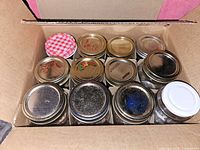 Twelve Bernardin Mason glass canning jars packed in a box, viewed from the top showing metal lids in silver and one red and white checkered lid.