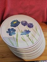 Stack of 12 plastic melamine plates with blue and purple flower design on a white background, displayed to show front of top plate.