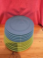 Stack of 6 blue melamine plates on top of 6 green melamine plates, arranged on wooden surface with pink fabric background.