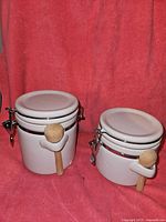 Two white ceramic jars side by side with their wooden scooping spoons attached to the side metal clamps.