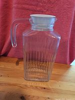 Front view of clear glass juice jug showing ribbed texture, handle, and white plastic lid on wooden table with red backdrop.