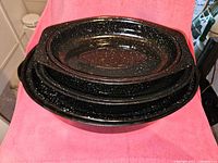 Stacked view of the three black enamelware roasting pans showing the lids on top, each pan varying in size with white speckled pattern on black enamel.