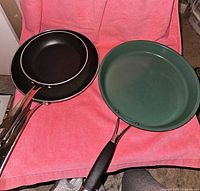 Three frying pans displayed on a pink towel: one green Paderno ceramic pan and two black Lagostina non-stick pans with metal handles.