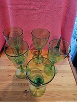 Six green translucent plastic wine glasses on a wooden surface with a red background. One glass is missing its base and placed inside another glass for display.