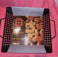 Top-down angled view of black perforated BBQ grill basket showing label with image of grilled food inside basket.