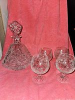 Full set of lead crystal brandy decanter and four snifter glasses placed in front of a red background showing design and number of items.