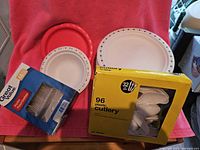 Lot contents including multiple sizes of white Chinet plates with blue floral border, red disposable bowls, unopened packs of plastic cutlery boxed in yellow and blue packaging.