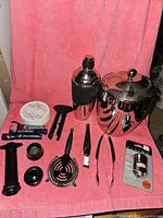 Full set of cocktail and wine accessories arranged on pink cloth backdrop showing ice bucket, shaker, strainer, tongs, lemon zester, wine openers, pump and coasters