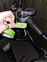Close-up of left handlebar grip showing brake lever, handle grip, and silver bell. Hand holding a green tape labeled 100 in front of bike frame.