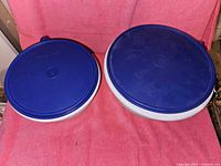 Two gray Tupperware stacking bowls with dark blue lids seen from above, showing the lids securely placed on top. The bowls are different sizes, approximately 12" and 10" diameter.