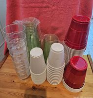 Photo showing stacked clear plastic wine glasses, turquoise, green, white, white styrofoam, and red disposable cups arranged on a wooden surface.