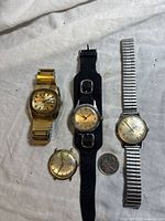 Front view of four mechanical wristwatches on white fabric, including a gold-colored watch, black leather strap watch, silver band watch, and small round watch.