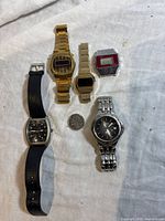 Five quartz wristwatches laid out on a white cloth. Includes two Bulova digital with gold-tone bands, one Timex digital, and two analog watches with silver-tone and black bands. A quarter is placed for scale.