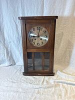 Front view of antique wooden wall clock with closed glass door, round gold-tone face with black numerals.