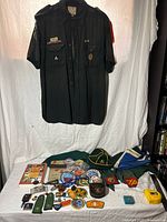 Photo showing full lot with both Scouts Canada vintage uniform shirts hanging and assortment of badges, patches, pins, hats, booklets, and belt buckles laid out on a table beneath them.