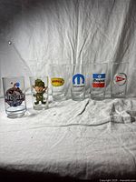Six vintage drinking glasses with automotive Mopar logos and two cartoon characters, shown six in a row on a white fabric background.