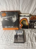 Four items displayed on fabric background: Three Bob Dylan books and one DVD case.