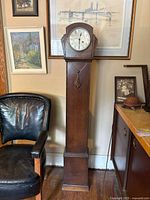 Full view of grandmother clock in room with furniture shows tall wooden cabinet and clock face at top.
