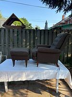 Recliner chair and matching ottoman placed on a white cloth-covered table outdoors against a wooden fence, showing side view of the furniture.