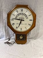 Front view of Schweppes Mixers electric advertising wall clock with octagonal wood frame, Roman numerals and pendulum window.