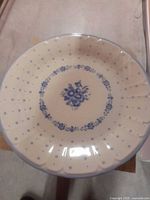 Top view of a ceramic plate featuring a blue floral vase design in the center surrounded by a ring of blue flowers and small blue dots on a cream colored surface with a scalloped edge.