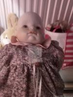 Close-up of an antique porcelain baby doll wearing a floral dress with pink collar and lace bib detail, shown with stuffed animals and a striped bag in background.