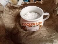 Top and side view of a ceramic soup cup with 'CHICKEN SOUP' text and vegetable illustrations. Cup is filled with white packing material.