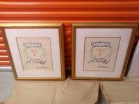 Two identical framed prints with colorful abstract faces, shown upright and side by side against an orange background.