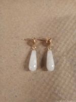 Photo showing white jade drop-shaped earrings with gold-tone posts on a beige textured background.
