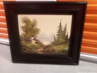 Framed landscape painting depicting a cabin near a stream among trees and greenery.