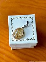 Pendant on decorative white box showing oval rutilated quartz set in gold bezel, visible rutile inclusions