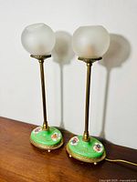 Pair of antique boudoir lamps with frosted globular glass shades and hand-painted floral porcelain bases with gilded brass accents shown standing side by side on wooden surface.