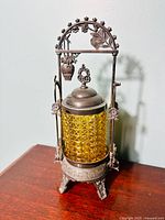 Front view of the antique pickle cruet showing amber colored patterned glass and decorative metal frame with bows and floral elements.