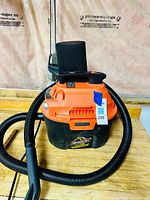 Front view of the orange and black Armor All wet/dry vacuum with hose attached, sitting on a wooden workbench.