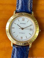 Front view of watch showing white dial, gold tone bezel, roman and stick hour markers, date window at 3 o'clock, and navy blue leather strap.